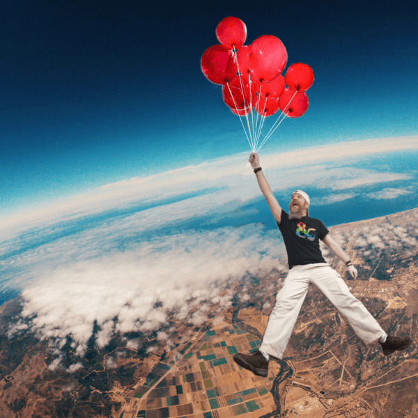 SteviL of Noble Knight Games is shown as a tall man in a Dungeons & Dragons logo tee, holding a bunch of red balloons by the string, while he floats happily miles above the earth