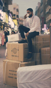 a man sitting on top of a pile of boxes in a busy market street,while sipping from a coffee cup