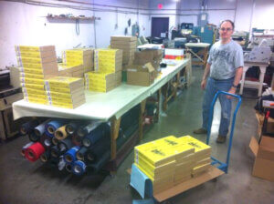 ian weir owner of red sash games at a print shop with stacks of copies of his wargame Yellow Jack