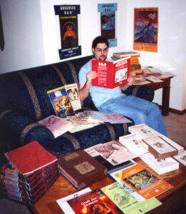 aaron leeder sitting on a sofa with a large collection of rare and out-of-print RPG books and modules