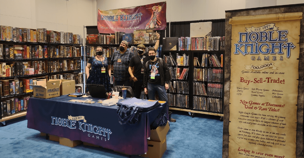 Noble Knight Games staff standing in a display booth surrounded by shelves of board games