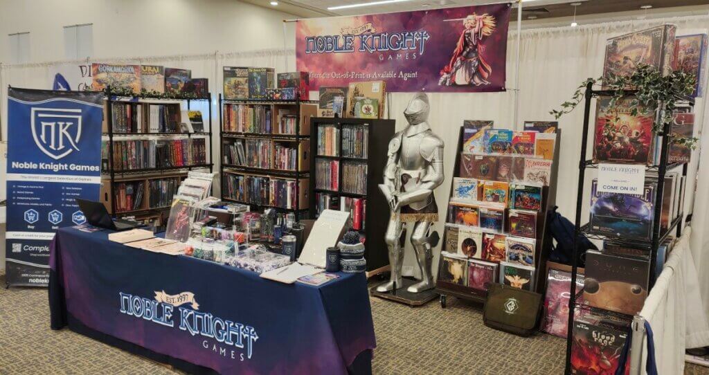 Photo of Noble Knight Games convention vendor booth with games, suit of armor, and Noble Knight Games banners